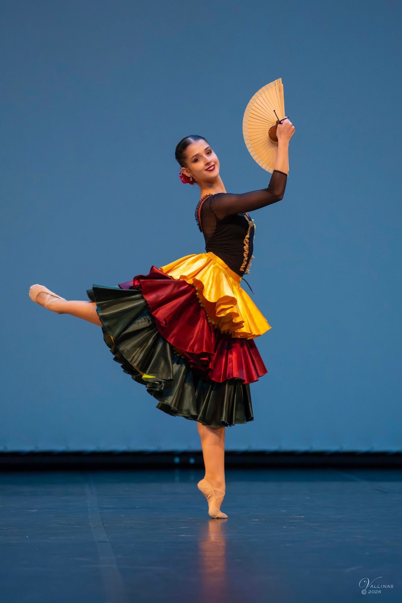 Bailarina de escuela bolera sonriendo en arabesque sobre puntas con abanico y falda de volantes de colores