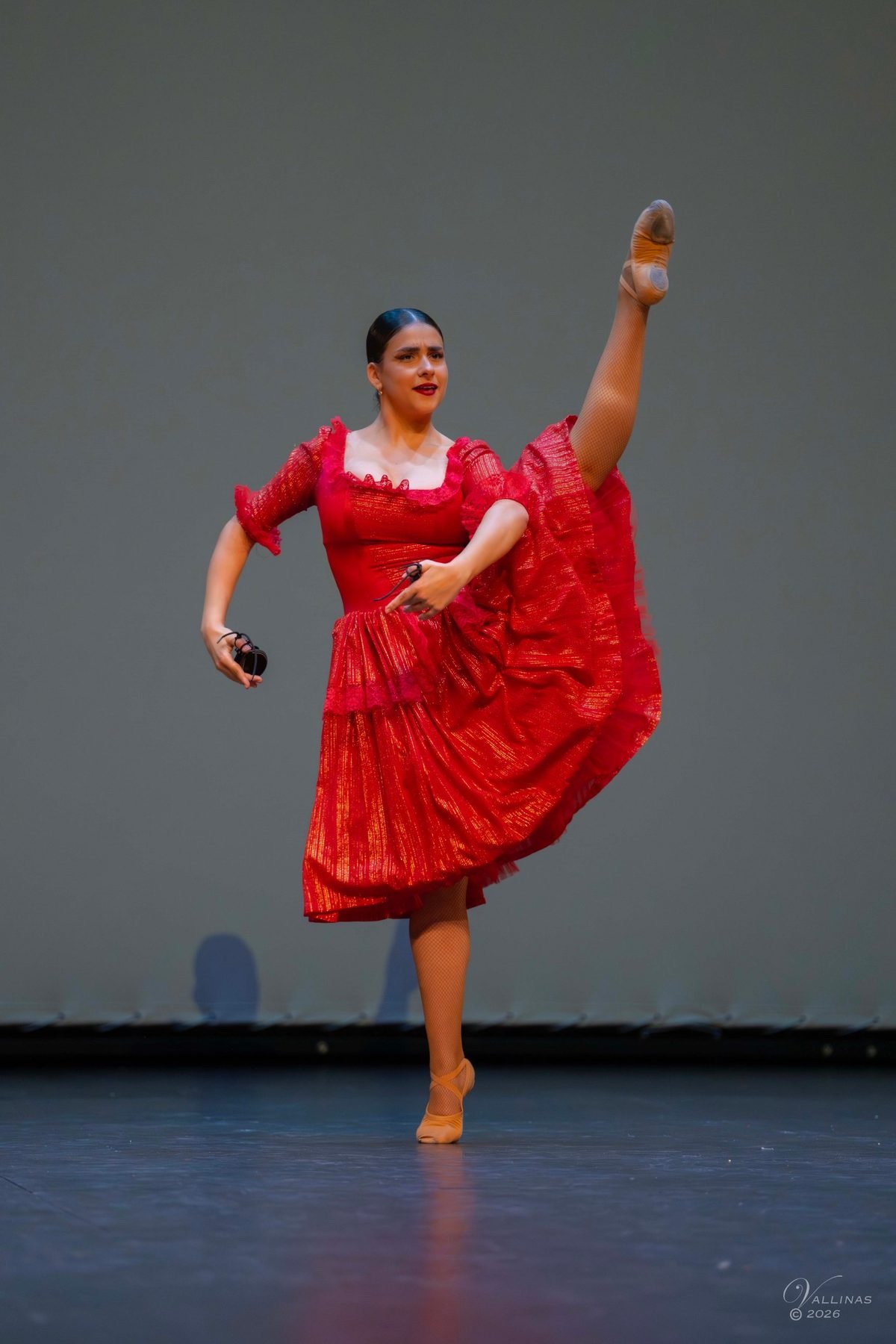 Bailarina de escuela bolera con vestido rojo y castañuelas, con la pierna extendida por encima de la cabeza en el escenario de Dance Magna 2026. Foto firmada por Jesús Vallinas